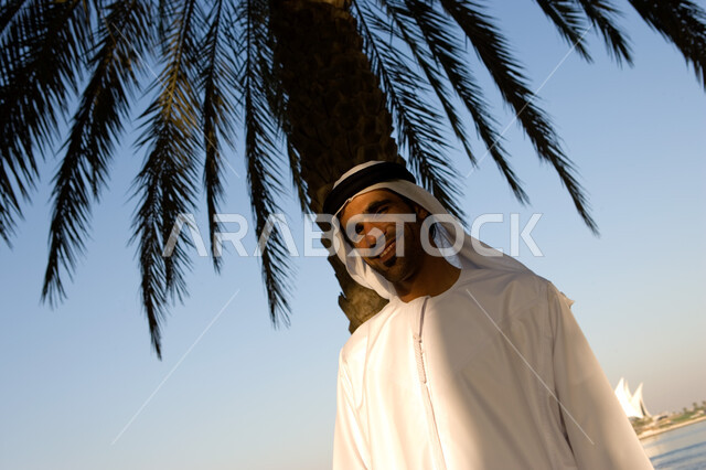 الاحتماء من حر الشمس تحت ظلال شجرة النخيل, صورة مقربة لرجل عربي خليجي إماراتي يرتدي الغترة والكندورة الإماراتية يقف على الكورنيش تحت شجرة النخيل بإيماءات تدل على السعادة, قضاء وقت الإجازة بالاستمتاع بالهواء الطلق, السياحة في الامارات العربية المتحدة