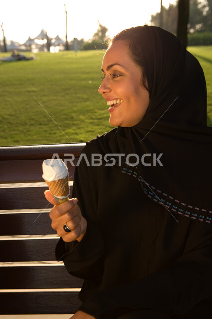 التخفيف من حر الصيف بتناول الآيس كريم, صورة مقربة لإمرأة عربية خليجية إماراتية مبتسمة ترتدي العباءة الإماراتية تجلس في الحديقة وتتناول المثلجات بإيماءات تدل على السعادة و الاستمتاع, قضاء أوقات الفراغ بالتنزه والترويح عن النفس , منتزهات وحدائق خضراء مميزة