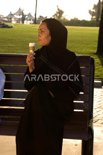 قضاء أوقات الفراغ بالتنزه والترويح عن النفس, التخفيف من حر الصيف بتناول الآيس كريم, صورة مقربة لإمرأة عربية خليجية إماراتية مبتسمة ترتدي العباءة الإماراتية تجلس في الحديقة وتتناول المثلجات بإيماءات تدل على السعادة و الاستمتاع, منتزهات وحدائق خضراء مميزة