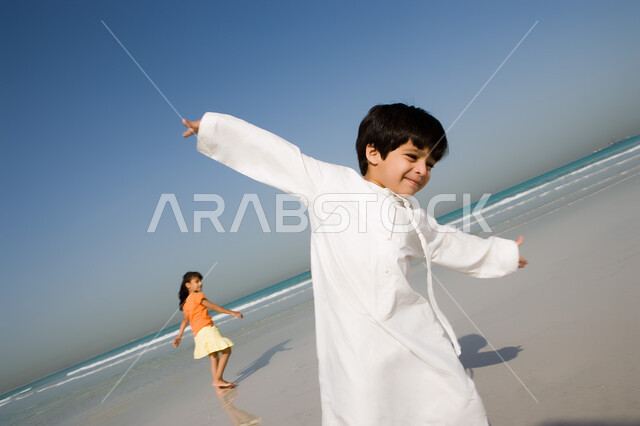 أجواء صيفية ممتعة, صورة مقربة لطفل عربي إمراتي خليجي مبتسم يقوم باللعب على شاطئ البحر رفقة أخته في رحلة صيفية, أنشطة ترفيهية عائلية في العطلة, الإستمتاع بالفعاليات مع الأخوة, منتجعات شاطئية في الإمارات