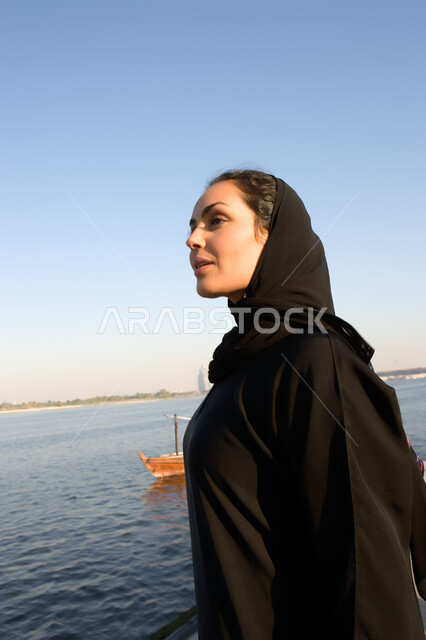 Leisure and entertainment and enjoying the summer vacation, spending vacation time enjoying the outdoors, tourism in the United Arab Emirates, a close-up of the side of an Arab Emirati woman wearing the Emirati abaya standing on the corniche with gestures