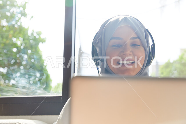 الترفيه بقضاء أوقات مسلية في أيام العطلة، القيام بمكالمة فيديو عبر الحاسوب المحمول، صورة مقربة لإمرأة عربيه إماراتية خليجية محجبة تجلس في غرفة المعيشة و ترتدي سماعات الرأس، التواصل مع الأهل و الأصدقاء عن بعد، إيماءات السعادة والاستمتاع باستخدام الانترنت