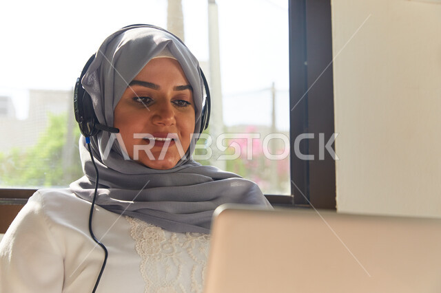 Entertaining by spending fun times during the holidays, making a video call via the laptop, a close-up of a veiled Emirati Arab woman sitting in the living room and wearing headphones, communicating with family and friends from a distance, gestures of hap