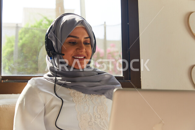 القيام بمكالمة فيديو عبر الحاسوب المحمول، الترفيه بقضاء أوقات مسلية في أيام العطلة، صورة مقربة لإمرأة عربيه إماراتية خليجية محجبة تجلس في غرفة المعيشة و ترتدي سماعات الرأس، التواصل مع الأهل و الأصدقاء عن بعد، إيماءات السعادة والاستمتاع باستخدام الانترنت