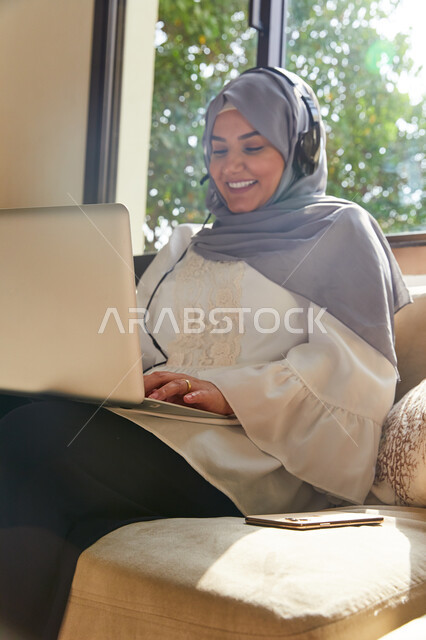 Communicating with friends and family from a distance, spending fun times on vacation days, making a video call via laptop, a close-up of a veiled Emirati Arab Gulf woman sitting in the living room and wearing headphones, gestures of happiness and enjoyin