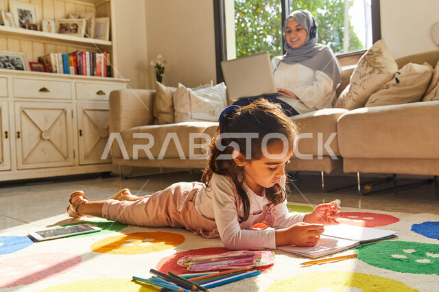 التسويق الإلكتروني في المنزل, تنمية القدرات والمهارات الفكرية للأطفال، ام عربية اماراتية خليجية محجبة تجلس في غرفة المعيشة  تضع سماعات الهيدفون وتقوم بالعمل على جهاز الحاسوب المحمول بجانب ابنتها، فتاة عربية اماراتية تقوم بالرسم والتلوين، العمل عن بعد