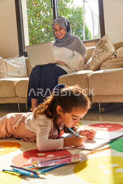 E-marketing at home, developing children's intellectual abilities and skills, a veiled Arab Emirati Gulf mother sitting in the living room putting on headphones and working on the laptop next to her daughter, an Arab Emirati girl drawing and coloring, wor