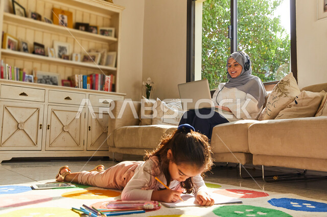 تنمية القدرات والمهارات الفكرية للأطفال، التسويق الإلكتروني في المنزل, ام عربية اماراتية خليجية محجبة تجلس في غرفة المعيشة  تضع سماعات الهيدفون وتقوم بالعمل على جهاز الحاسوب المحمول بجانب ابنتها، فتاة عربية اماراتية تقوم بالرسم والتلوين، العمل عن بعد