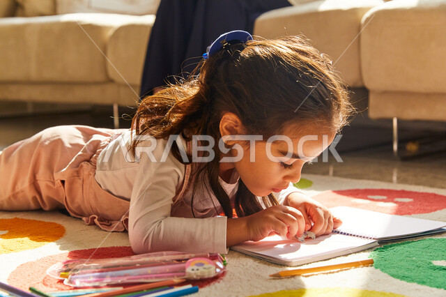 أنشطة ترفيهية فنية ابداعية، صورة مقربة لطفلة عربية خليجية إماراتية تجلس في غرفة المعيشة وتبدع بتلوين الصور بجانب والدتها، أدوات ومعدات التلوين، التعلم والابداع وتنمية المهارات، قضاء اوقات ممتعة بتلوين الرسومات، ايماءات وجه تدل على الاندماج و التركيز 