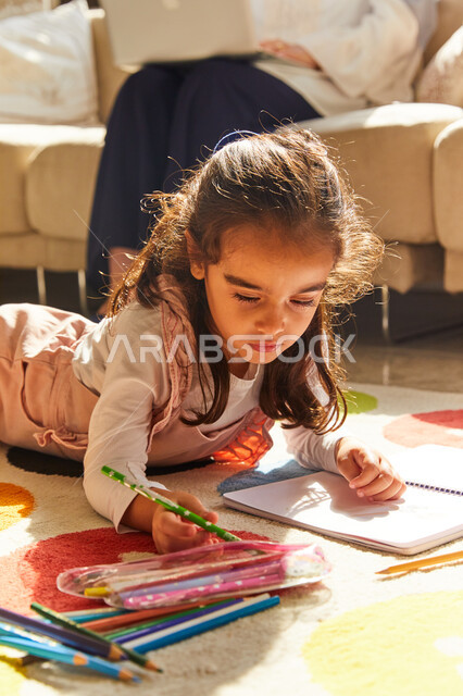 Creative artistic entertainment activities, a close-up of an Arab Gulf Emirati girl sitting in the living room and being creative by coloring pictures next to her mother, coloring tools and equipment, learning, creativity and skill development, spending f