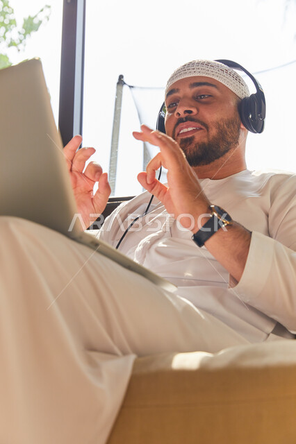 التواصل مع الأهل والرفاق، استخدام تقنية الهيدفون، صورة مقربه لرجل عربي اماراتي خليجي مبتسم يرتدي الطاقية البيضاء والكندورة وسماعه الرأس الصوتية، التسويق الالكتروني من المنزل، إجراء مكالمة فيديو عبر الحاسوب المحمول, إيماءات تدل على التأكيد