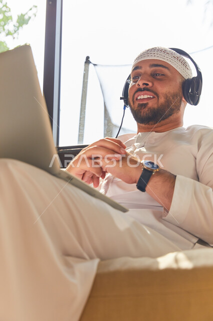التواصل مع الأهل والرفاق، استخدام تقنية الهيدفون، صورة مقربه لرجل عربي اماراتي خليجي مبتسم يرتدي الطاقية البيضاء والكندورة وسماعه الرأس الصوتية، التسويق الالكتروني من المنزل، إجراء مكالمة فيديو عبر الحاسوب المحمول