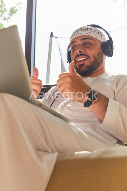 استخدام تقنية الهيدفون، التواصل مع الأهل والرفاق، صورة مقربه لرجل عربي اماراتي خليجي مبتسم يرتدي الطاقية البيضاء والكندورة وسماعه الرأس الصوتية، التسويق الالكتروني من المنزل، إجراء مكالمة فيديو عبر الحاسوب المحمول, إيماءات تدل على التأكيد