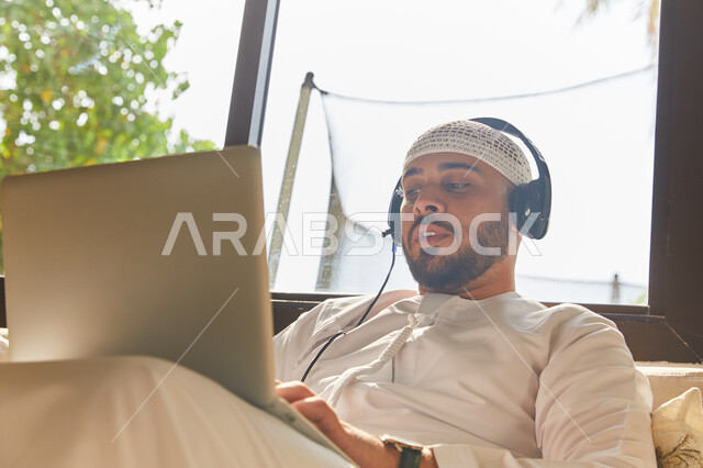 إجراء مكالمة فيديو عبر الحاسوب المحمول, التواصل مع الأهل والرفاق، استخدام تقنية الهيدفون، صورة مقربه لرجل عربي اماراتي خليجي مبتسم يرتدي الطاقية البيضاء والكندورة وسماعه الرأس الصوتية، التسويق الالكتروني من المنزل