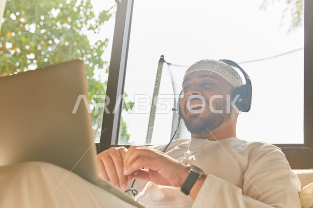 إجراء مكالمة فيديو عبر الحاسوب المحمول, التواصل مع الأهل والرفاق، استخدام تقنية الهيدفون، صورة مقربه لرجل عربي اماراتي خليجي مبتسم يرتدي الطاقية البيضاء والكندورة وسماعه الرأس الصوتية، التسويق الالكتروني من المنزل