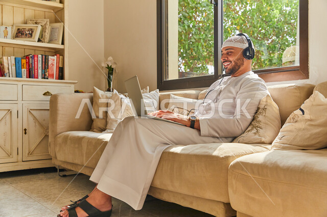 تصفج مواقع التواصل الاجتماعي, إيماءات الاستمتاع بالتواصل مع الأهل والرفاق، استخدام تقنية الهيدفون، صورة من الجانب لرجل عربي اماراتي خليجي مبتسم يرتدي الطاقية البيضاء والكندورة وسماعه الرأس الصوتية، إجراء مكالمة فيديو عبر الحاسوب المحمول