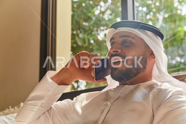 الاستمتاع بتبادل الأحاديث و الأخبار، إجراء مكالمة هاتفية صوتية عبر الجوال، صورة مقربة لرجل عربي اماراتي خليجي يرتدي الكندورة والغترة الاماراتية يجلس في غرفة المعيشة ويتحدث عبر الهاتف المحمول، إيماءات السعادة بالتواصل مع الأصدقاء والأقارب
