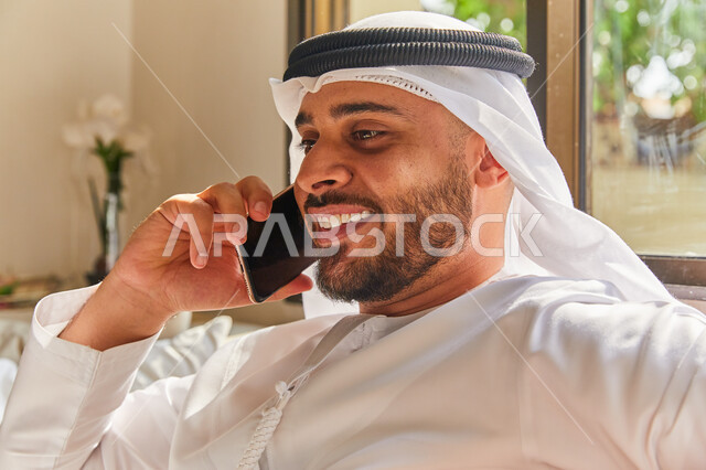 Enjoying the exchange of conversations and news, making a voice phone call via mobile phone, a close-up of an Arab Emirati Gulf man wearing the Emirati kandora and ghutra sitting in the living room and talking on the mobile phone, gestures of happiness by