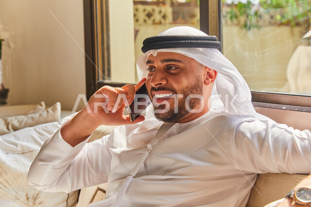 إجراء مكالمة هاتفية صوتية عبر الجوال، الاستمتاع بتبادل الأحاديث و الأخبار، صورة مقربة لرجل عربي اماراتي خليجي يرتدي الكندورة والغترة الاماراتية يجلس في غرفة المعيشة ويتحدث عبر الهاتف المحمول، إيماءات السعادة بالتواصل مع الأصدقاء والأقارب
