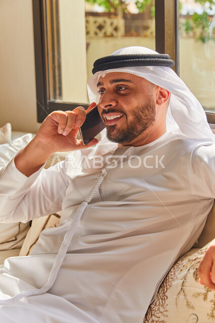 إجراء مكالمة هاتفية صوتية عبر الجوال، الاستمتاع بتبادل الأحاديث و الأخبار، صورة مقربة لرجل عربي اماراتي خليجي يرتدي الكندورة والغترة الاماراتية يجلس في غرفة المعيشة ويتحدث عبر الهاتف المحمول، إيماءات السعادة بالتواصل مع الأصدقاء والأقارب