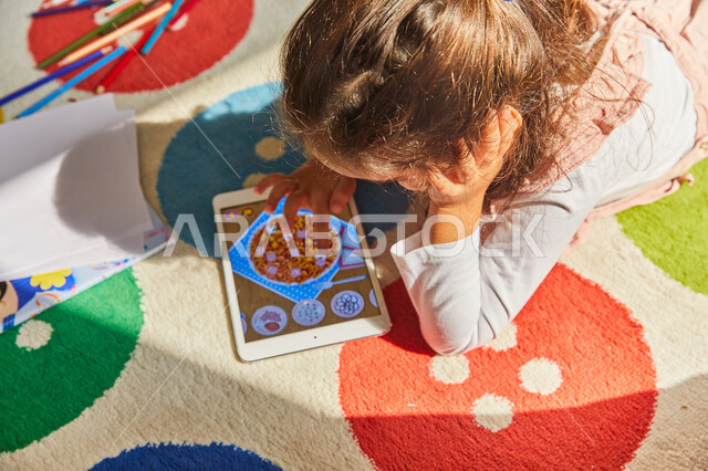 Exploiting leisure time with fun recreational activities, a close-up of an Arab Gulf Emirati girl sitting on the ground next to drawing tools and using a tablet, developing children's mental and motor skills, caring for children and taking interest in use