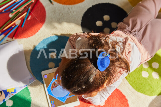استغلال أوقات الفراغ بالأنشطة الترفيهية الممتعة, صورة مقربة لفتاة عربية خليجية إماراتية تجلس على الأرض بجانب أدوات الرسم وتقوم باستخدام الجهاز اللوحي, تنمية مهارات الأولاد الذهنية والحركية, رعاية الأبناء والاهتمام بالهوايات المفيدة 