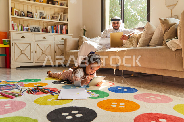 التسلية والترفيه بوقت الفراغ، التسوق الإلكتروني من المنزل, أب عربي امراتي خليجي يرتدي الزي الإماراتي يجلس في غرفة المعيشة ويستخدم التابلت، فتاة عربية اماراتية تجلس على الأرض و تقوم بالعب على الجهاز اللوحي ، العمل عن بعد في المنزل رفقة الأبناء