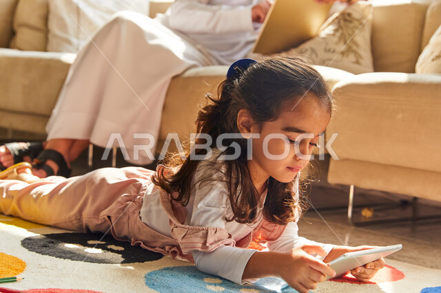 التسوق الإلكتروني من المنزل, التسلية والترفيه بوقت الفراغ، أب عربي امراتي خليجي يرتدي الزي الإماراتي يجلس في غرفة المعيشة ويستخدم التابلت، صورة مقربة لفتاة عربية اماراتية تجلس على الأرض و تقوم بالعب على الجهاز اللوحي ، العمل عن بعد في المنزل رفقة الأبناء