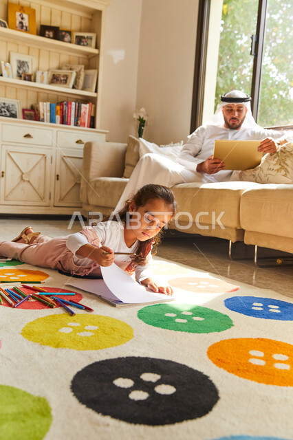 التعلم وتنمية المهارات الفنية للأطفال، أدوات ومعدات الرسم و التلوين، فتاة عربية خليجية إماراتية تجلس على الأرض وتقوم بالرسم بالألوان، أب إماراتي يرتدي الزي الإماراتي يجلس في غرفة المعيشة مع ابنته و يحمل التابلت, تصفح وسائل التواصل الاجتماعي