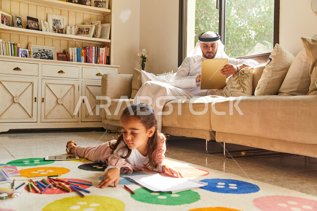 Learning and developing children's artistic skills, drawing and coloring tools and equipment, an Emirati Arab Gulf girl sitting on the floor and drawing in colors, an Emirati father wearing Emirati clothes sitting in the living room with his daughter and 