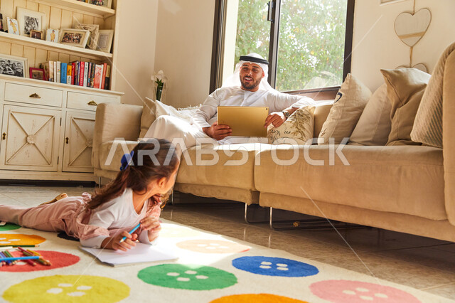 Drawing and coloring tools and equipment, learning and developing artistic skills for children, an Emirati Arab Gulf girl sitting on the floor and drawing in colors, an Emirati father wearing Emirati clothes holding a tablet and sitting in the living room