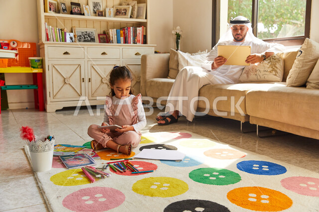 قضاء الوقت بتعلم  المهارات الفنية، أدوات ومعدات الرسم و التلوين، فتاة عربية خليجية إماراتية تجلس على الأرض وتقوم بالرسم بالألوان، أب إماراتي يرتدي الزي الإماراتي يجلس في غرفة المعيشة مع ابنته و يحمل التابلت, تصفح وسائل التواصل الاجتماعي