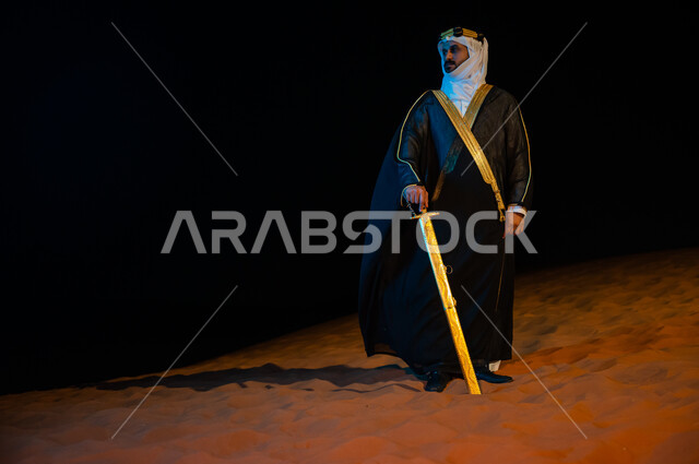 مفهوم الحراسه والمراقبه ليلا، رجل عربي خليجي سعودي يرتدي الغتره والبشت ويحمل بيده سيف في الخارج وقت الليل، الوقوف باعتزاز وثقة بدولة السعودية, التامل والنظر الى شيء ما, خلفية الطبيعه الصحراوية، صورة الجسم بالطول الكامل