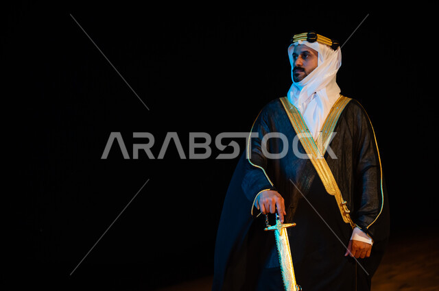 Standing with pride and confidence in the state of Saudi Arabia, wearing the traditional folk dress of the eastern region, a close-up of a Saudi Arabian Gulf man wearing a ghutra and a bisht and holding a sword in his hand outside at night time, contempla