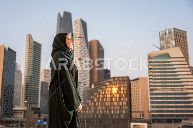 التجول واستنشاق الهواء النقي في الخارج, الفخر والاعتزاز بالتقدم المعماري في السعودية، النمو والتطور العمراني في مدينة الرياض، صورة من الجانب لسيدة عربية خليجية سعودية ترتدي العباءة والحجاب تنظر الى شيء ما في وقت النهار، خلفية ابراج الملك عبدلله الماليه