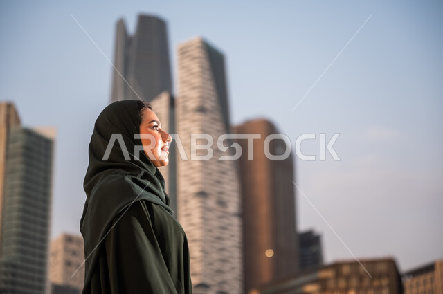 الاعتزاز والفخر بالتقدم المعماري في السعودية، النمو والتطور العمراني في مدينة الرياض، التجول واستنشاق الهواء النقي في الخارج، صورة مقربة لإمرأة عربية خليجية سعودية ترتدي العباءة والحجاب تنظر الى شيء ما في وقت النهار، خلفية ابراج الملك عبدلله الماليه