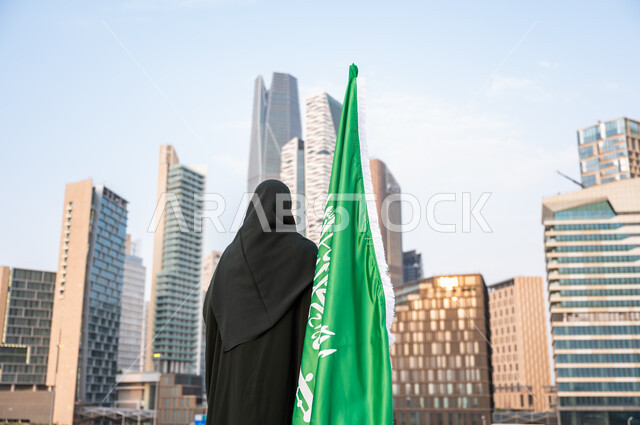 أبراج مدينة الرياض في وقت النهار, الاحتفال بتوحيد المملكة, ذكرى العلم السعودي 11 مارس, إحياء اليوم الوطني 23 سبتمبر, صورة من الخلف لامرأة عربية خليجية سعودية محجبة ترتدي العباءة السوداء تقف بجانب العلم السعودي