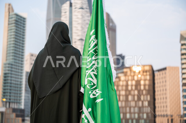 الاحتفال بتوحيد المملكة 23 سبتمبر, ذكرى العلم 11 مارس, إحياء اليوم الوطني السعودي, صورة من الخلف لامرأة عربية خليجية سعودية محجبة ترتدي العباءة السوداء تقف بجانب العلم السعودي, خلفية أبراج مدينة الرياض