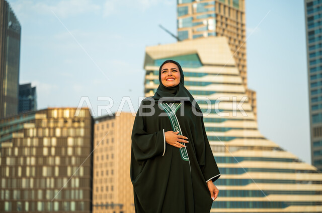 الاستمتاع والتجول في وقت النهار, مفهوم الإنشاءات والهندسة المعمارية, قضاء وقت ممتع في المدينة المالية, شابه عربية سعودية خليجية مبتسمة ترتدي العباءة والحجاب تنظر إلى شيء ما, جولة سياحية ترفيهيه في مدينة الرياض