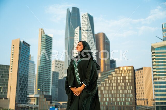 قضاء وقت ممتع في المدينة المالية, مفهوم الإنشاءات والهندسة المعمارية, شابه عربية سعودية خليجية مبتسمة ترتدي العباءة والحجاب تنظر إلى شيء ما, الاستمتاع والتجول في وقت النهار, جولة سياحية ترفيهيه في مدينة الرياض