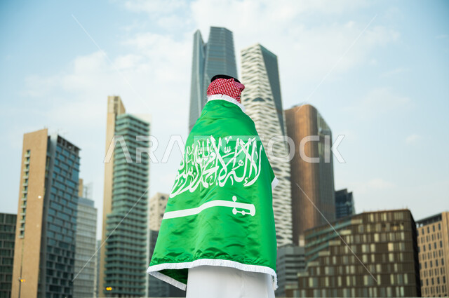 ارتداء الزي التقليدي السعودي بيوم العلم 11 مارس, إظهار الحب والولاء للدولة في اليوم الوطني 23 سبتمبر, صورة من الخلف لرجل عربي سعودي خليجي يرتدي علم المملكة بفخر وعز, الاحتفالات بذكرى العيد الوطني, حب الوطن والانتماء إليه, خلفية أبراج كافد المالية