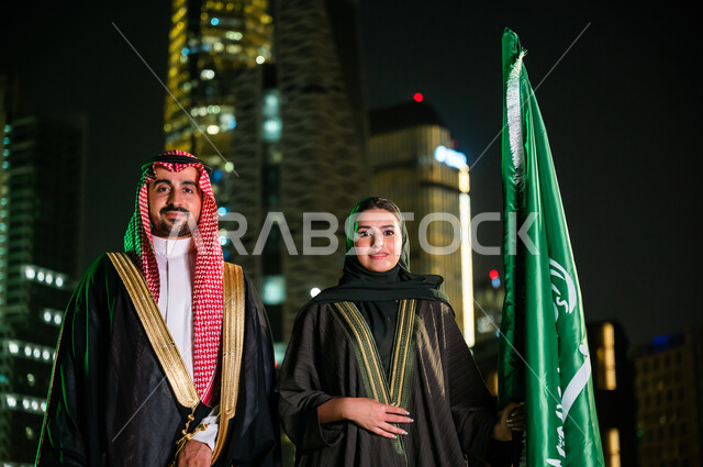 التعبير عن الحب والولاء للسعودية, العيد الوطني 8 ربيع الأول، الاحتفال بذكرى اليوم الوطني ، يوم العلم السعودي 11 مارس، زوجان عربيان سعوديان خليجيان يقفان بجانب العلم بثقة بإيماءات الفخر والاعتزاز بالوطن، خلفية أبراج وناطحات سحاب مدينة الرياض المضاءة ليلا.