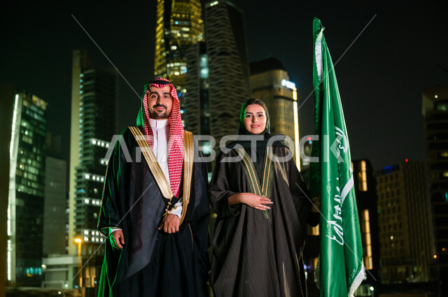 التعبير عن الحب والولاء للسعودية, العيد الوطني 8 ربيع الأول، الاحتفال بذكرى اليوم الوطني ، يوم العلم السعودي 11 مارس، زوجان عربيان سعوديان خليجيان يقفان بجانب العلم بثقة بإيماءات الفخر والاعتزاز بالوطن، خلفية أبراج وناطحات سحاب مدينة الرياض المضاءة ليلا.