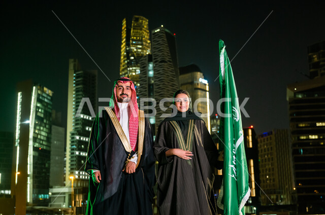 التعبير عن الحب والولاء للسعودية, العيد الوطني 8 ربيع الأول، الاحتفال بذكرى اليوم الوطني ، يوم العلم السعودي 11 مارس، زوجان عربيان سعوديان خليجيان يقفان بجانب العلم بثقة بإيماءات الفخر والاعتزاز بالوطن، خلفية أبراج وناطحات سحاب مدينة الرياض المضاءة ليلا.