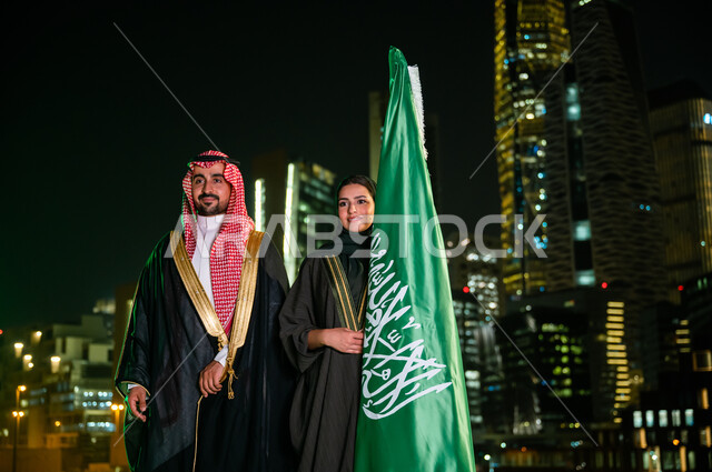 Celebrating the anniversary of the National Day, Saudi Flag Day, March 11, expressing love and loyalty to Saudi Arabia, National Day, Rabi’ al-Awwal 8, a Saudi-Gulf Arab couple standing next to the flag confidently with gestures of pride and pride in the homeland, the background of the towers and skyscrapers of the city of Riyadh illuminated at night.