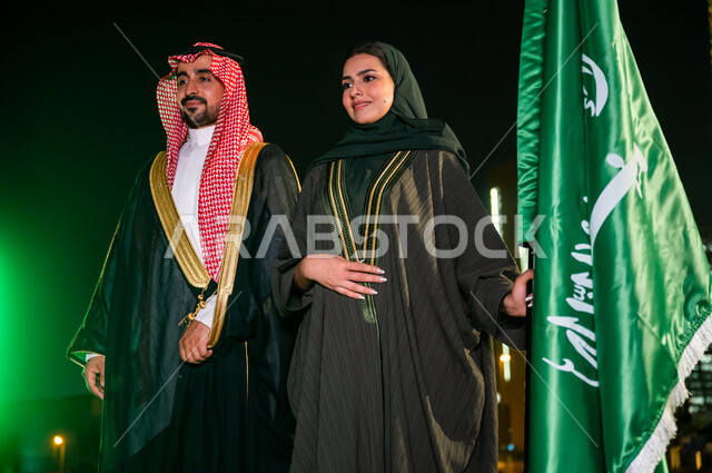 Celebrating the anniversary of the National Day, Saudi Flag Day, March 11, expressing love and loyalty to Saudi Arabia, National Day, Rabi’ al-Awwal 8, a Saudi-Gulf Arab couple standing next to the flag confidently with gestures of pride and pride in the homeland, the background of the towers and skyscrapers of the city of Riyadh illuminated at night.