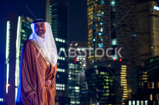 الوقوف بفخر واعتزاز بالدولة السعودية, الاستمتاع والخروج في الهواء الطلق, رجل عربي خليجي سعودي يرتدي البشت والغترة, ارتداء الأزياء الشعبية التقليدية، خلفية أبراج وناطحات سحاب مدينة الرياض المضاءة في وقت الليل.