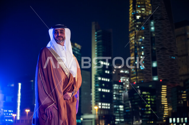 الوقوف بفخر واعتزاز بالدولة السعودية, الاستمتاع والخروج في الهواء الطلق, رجل عربي خليجي سعودي يرتدي البشت والغترة, ارتداء الأزياء الشعبية التقليدية، خلفية أبراج وناطحات سحاب مدينة الرياض المضاءة في وقت الليل.
