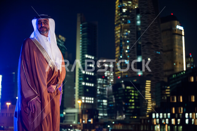الوقوف بفخر واعتزاز بالدولة السعودية, الاستمتاع والخروج في الهواء الطلق, رجل عربي خليجي سعودي يرتدي البشت والغترة, ارتداء الأزياء الشعبية التقليدية، خلفية أبراج وناطحات سحاب مدينة الرياض المضاءة في وقت الليل.
