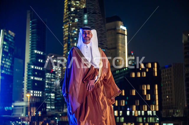 الفخر والاعتزاز بالدولة السعودية, ارتداء الأزياء الشعبية التقليدية، الوقوف بثقة والنظر إلى الكاميرا، الاستمتاع والخروج في الهواء الطلق, رجل عربي خليجي سعودي يرتدي البشت والغترة, خلفية أبراج وناطحات سحاب مدينة الرياض المضاءة في وقت الليل.